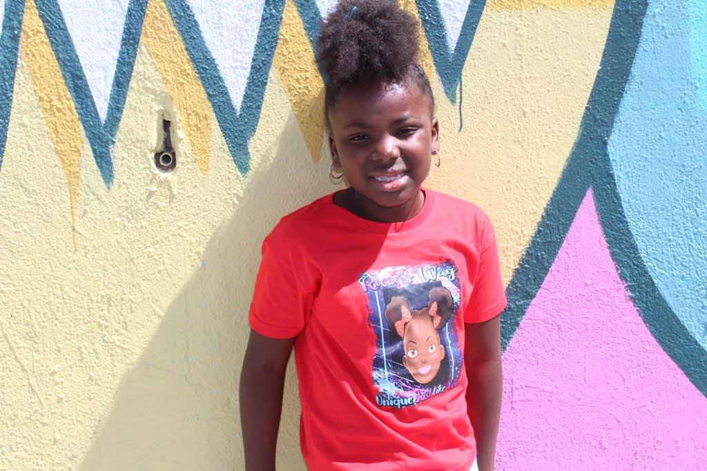 Little girl standing next to wall with Riley's Way t-shirt